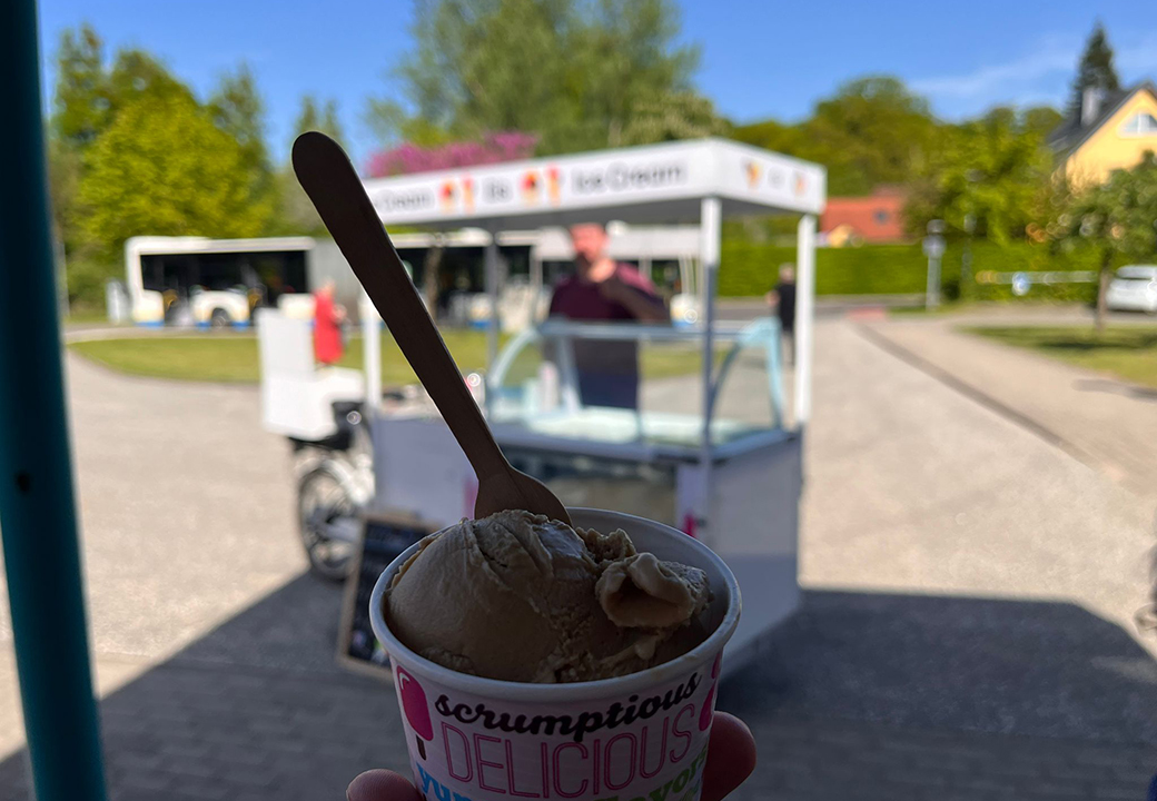 Eine Portion Eis in einem Becher im Vordergrund, mit einem Eisstand im Hintergrund.