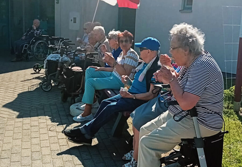 Eine Gruppe &auml;lterer Menschen sitzt auf einer Parkbank im Freien, teils mit Rollatoren oder Rollst&uuml;hlen, und genie&szlig;t Eiscreme.