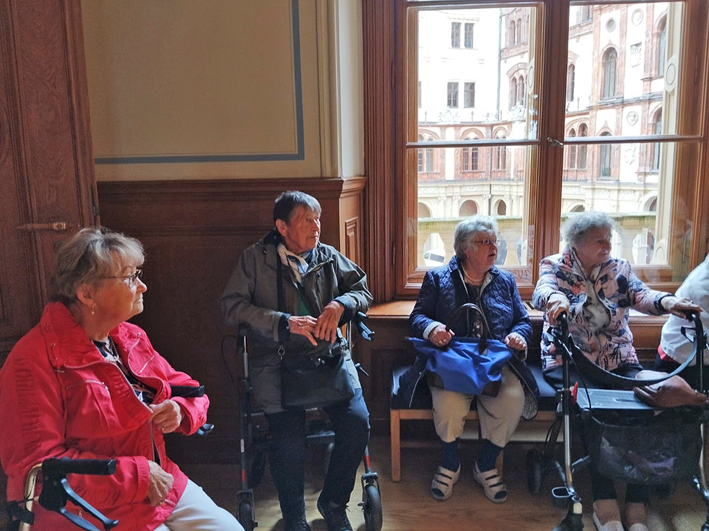 Vier ältere Frauen sitzen mit Rollatoren in einem Raum mit Fenster, durch das eine historische Fassade sichtbar ist.