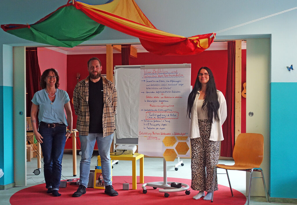 Drei Menschen stehen vor einem Flipchart in einem Raum mit buntem Dekor.