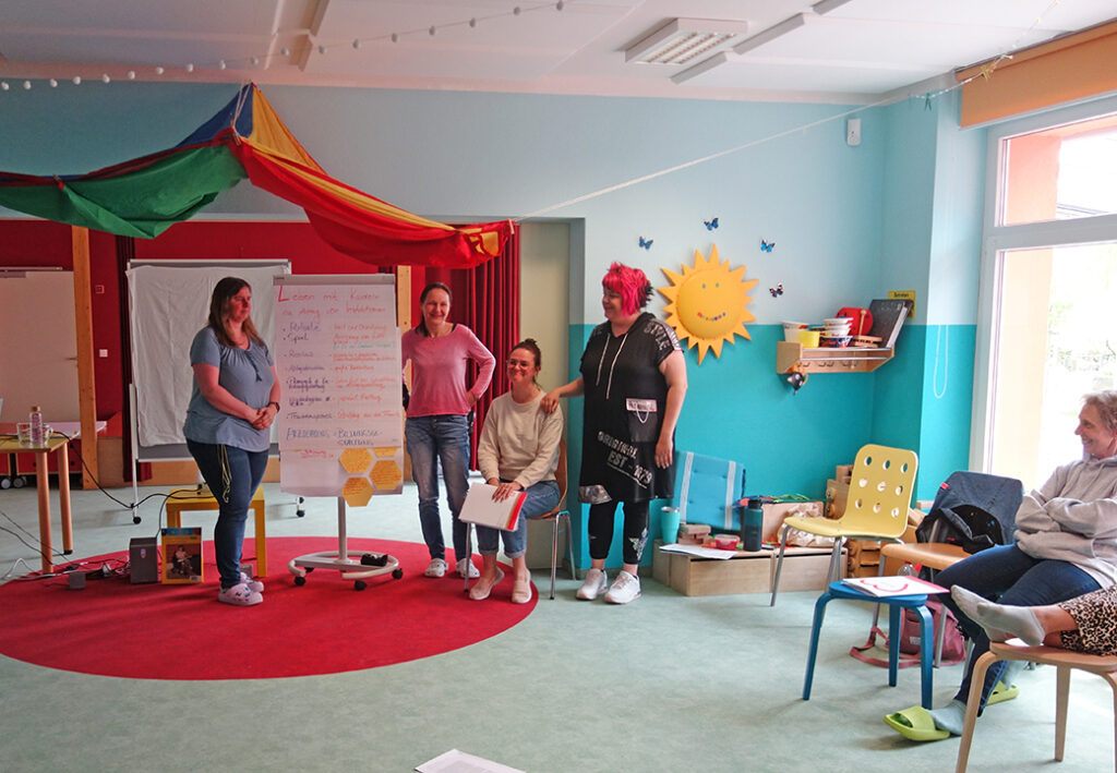 Eine Gruppe von Frauen steht und sitzt in einem bunt dekorierten Raum mit einem Flipchart in der Mitte.