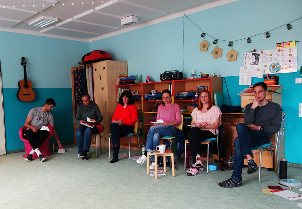 Eine Gruppe von sechs Personen sitzt in einem hellen Raum mit einer Gitarre und bunten Regalen.