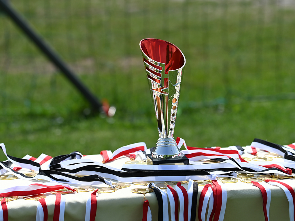 Ein silberner Pokal mit rotem Innenbereich steht auf einem Tisch, umgeben von roten, weißen und schwarzen Medaillen auf Bändern.
