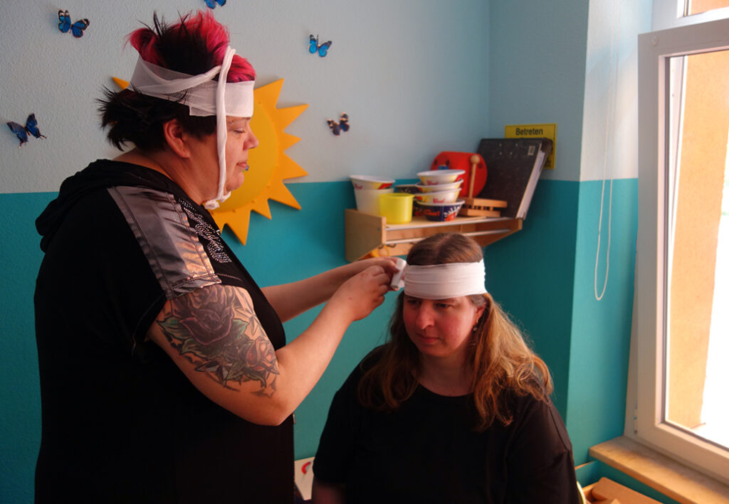 Eine Frau mit pinken Haaren verbindet den Kopf einer sitzenden Frau, die dunkelblonde Haare hat.