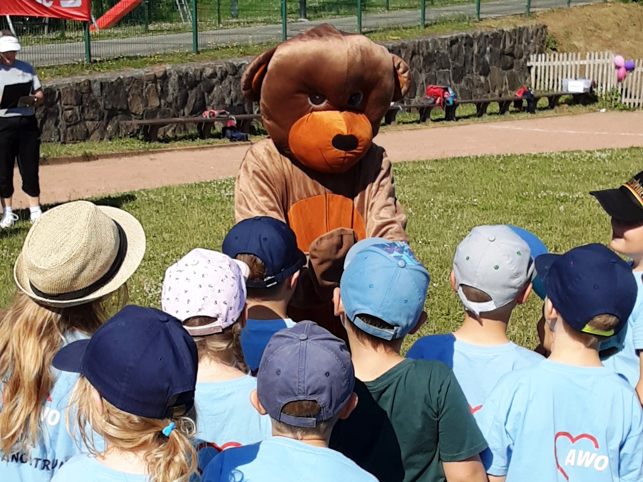 Gruppe von Kindern steht vor Person im Bärenkostüm auf einem Sportfeld