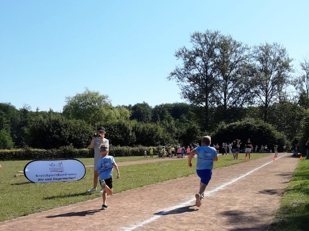 Kinder laufen ein Rennen auf einer Laufbahn im Freien. Ein Erwachsener steht daneben und h&auml;lt eine Kamera.