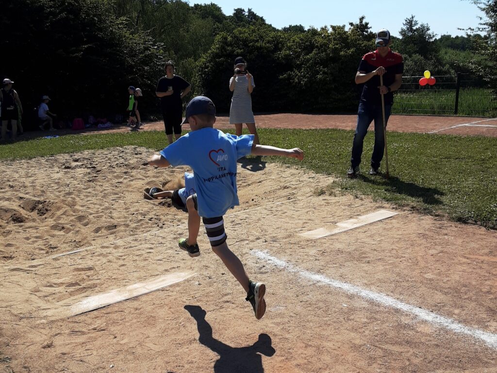 Ein Kind im blauen AWO-T-Shirt beim Weitsprung auf einem Sportplatz.
