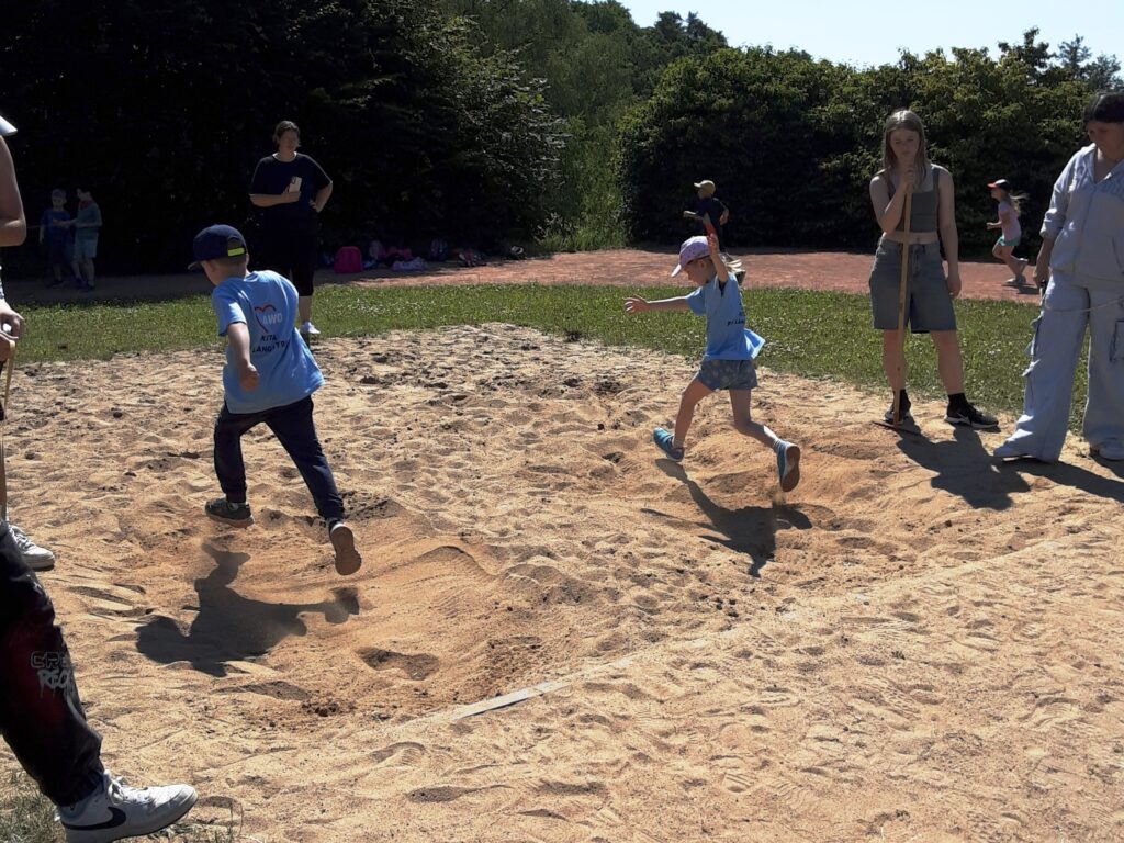 Zwei Kinder springen gleichzeitig in eine Sandgrube, w&auml;hrend Erwachsene zuschauen