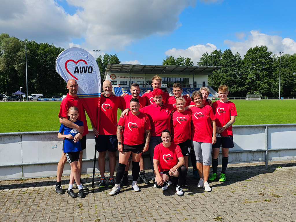 Eine Gruppe von Menschen steht lächelnd auf einem Sportplatz vor einer Tribüne. Sie tragen rote T-Shirts mit dem AWO-Logo.