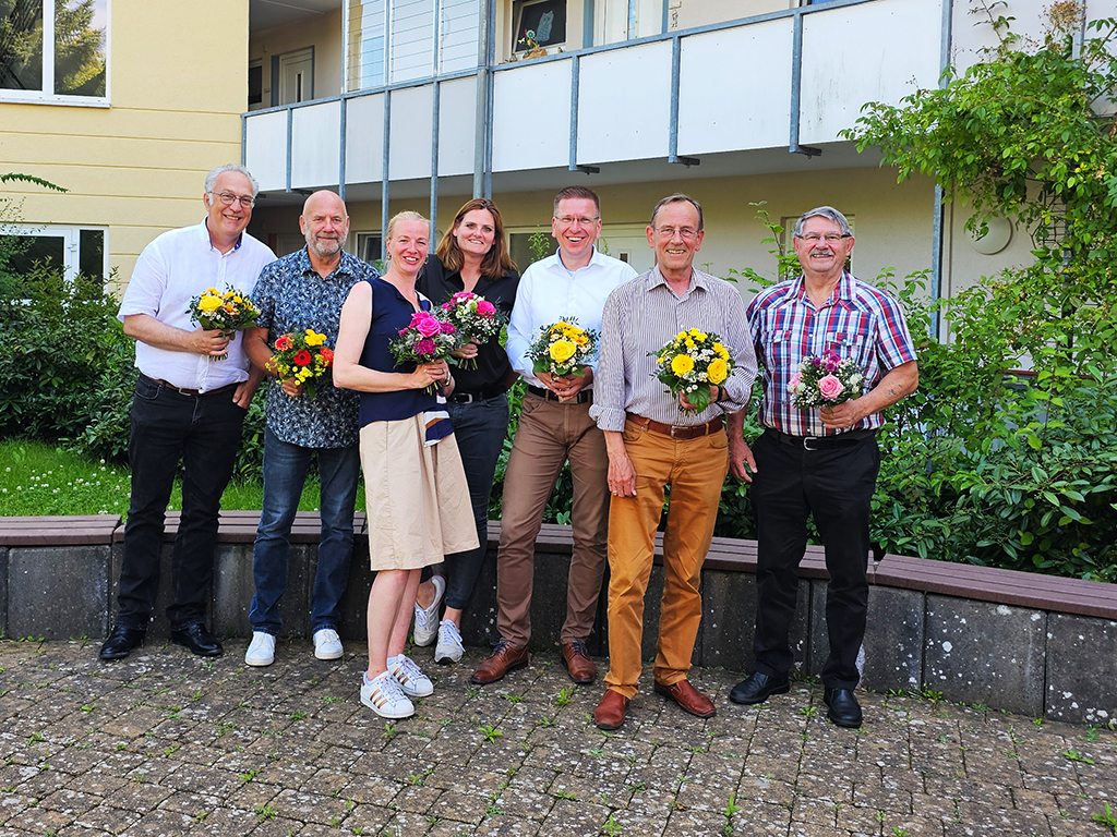 Eine Gruppe von sieben Menschen hält Blumensträuße in den Händen und steht vor einem Gebäude.