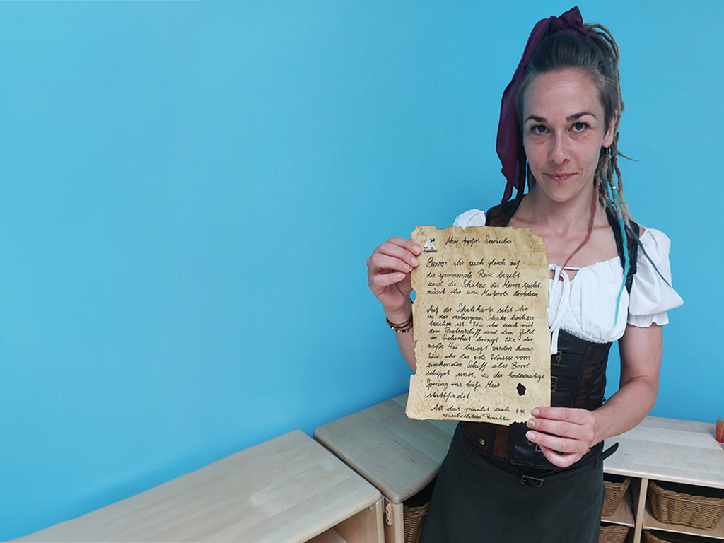 Frau in einem Piratenkostüm hält eine alt aussehende Schriftrolle vor einer blauen Wand.