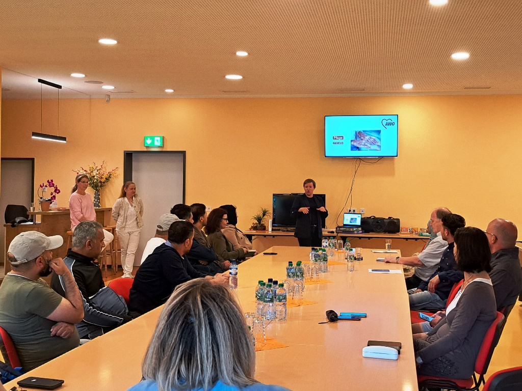 Menschen sitzen um einen Konferenztisch und hören einem Vortrag zu.