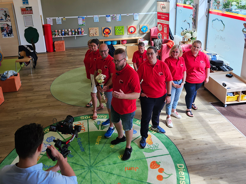 Eine Gruppe von Menschen in roten T-Shirts steht in einer Linie in einem Raum, während sie von einer Person mit einer Kamera gefilmt werden.