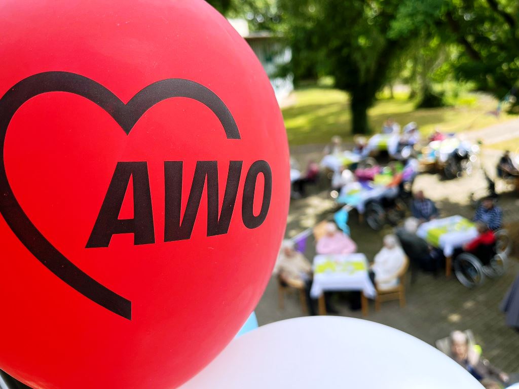 Ein roter Luftballon mit dem Logo der AWO im Vordergrund, im Hintergrund sind unscharf Menschen an Tischen im Freien zu sehen.