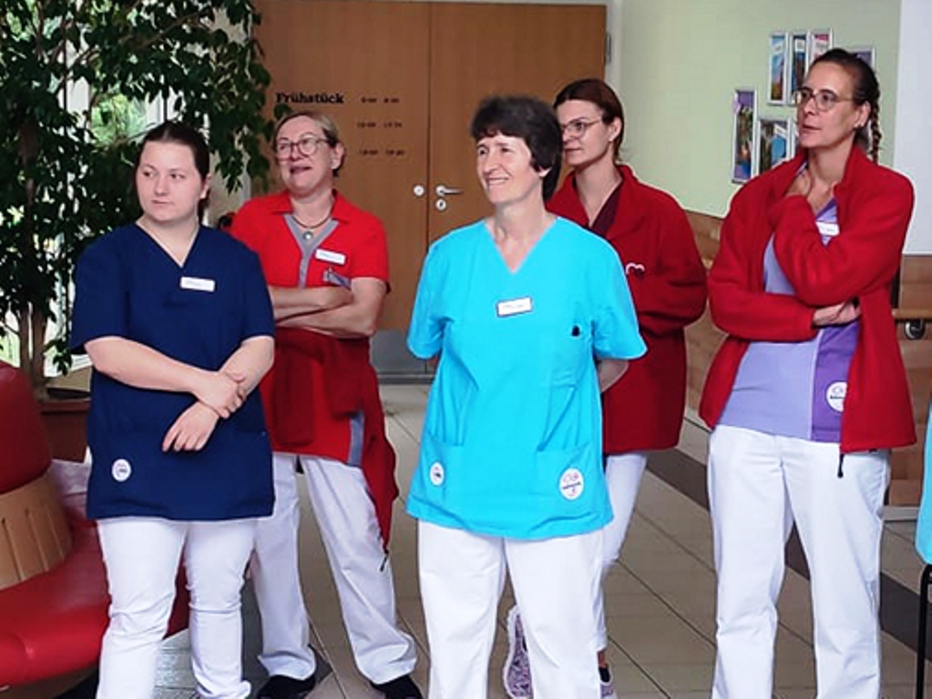 Eine Gruppe von Mitarbeitern in farbigen Arbeitskleidungen, darunter rote, blaue und violette Oberteile, steht in einem Innenbereich.