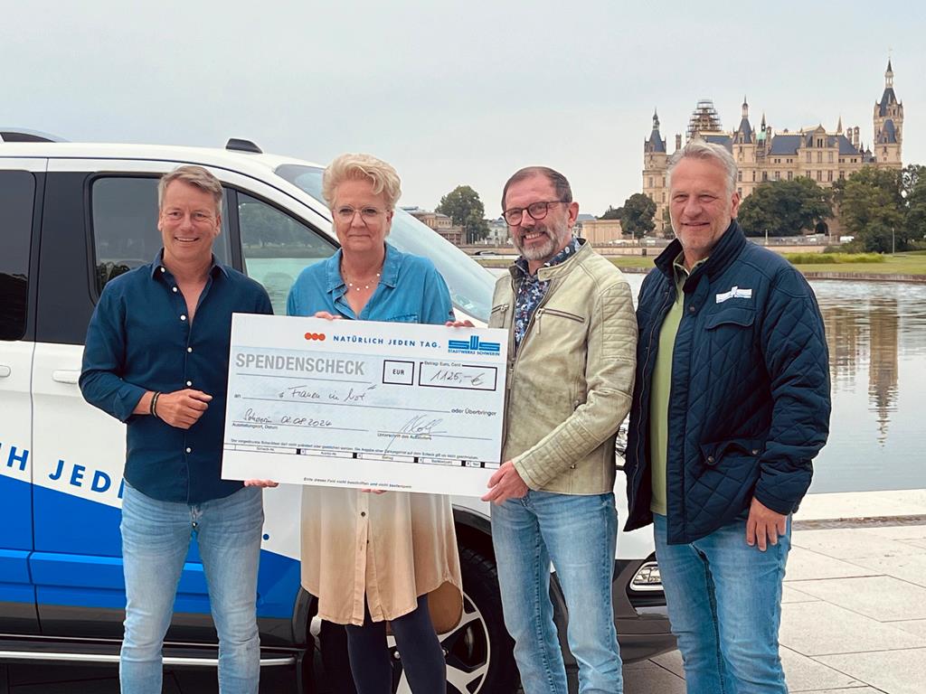 Vier Personen stehen draußen und halten einen großen Spendenscheck in den Händen. Im Hintergrund ist ein Auto und ein Schloss zu sehen.