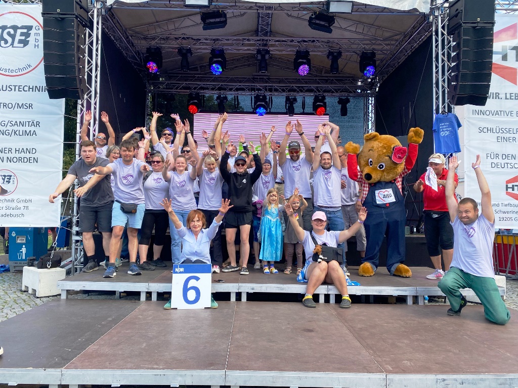 Gruppenfoto auf einer Bühne mit Menschen in lila T-Shirts und einem Maskottchen in Bärenkostüm