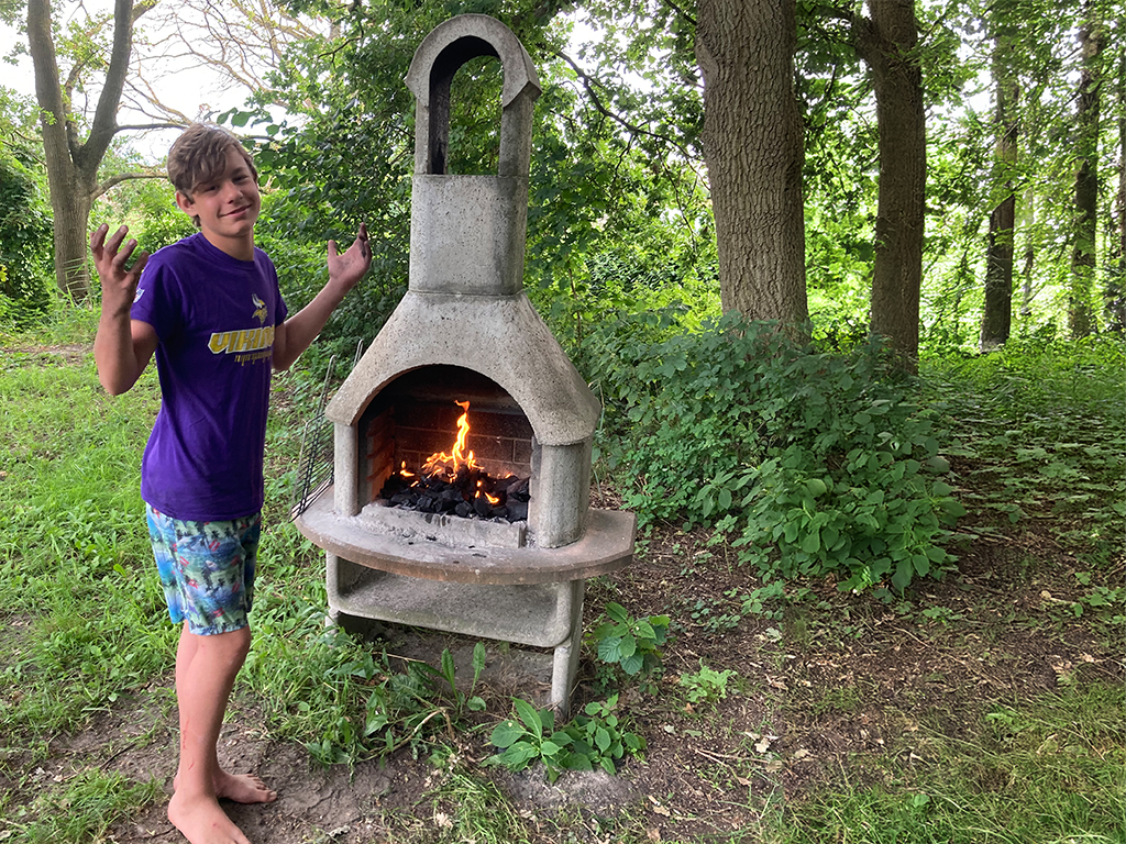 Ein Junge steht neben einem brennenden Kamin im Freien.