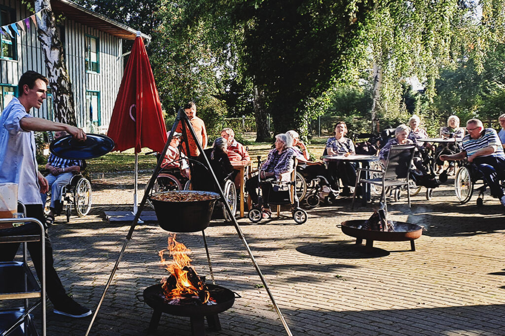 Gruppe von Menschen, einige in Rollst&uuml;hlen, bei einem Outdoor-Essen mit einem gro&szlig;en Kessel &uuml;ber einem offenen Feuer.