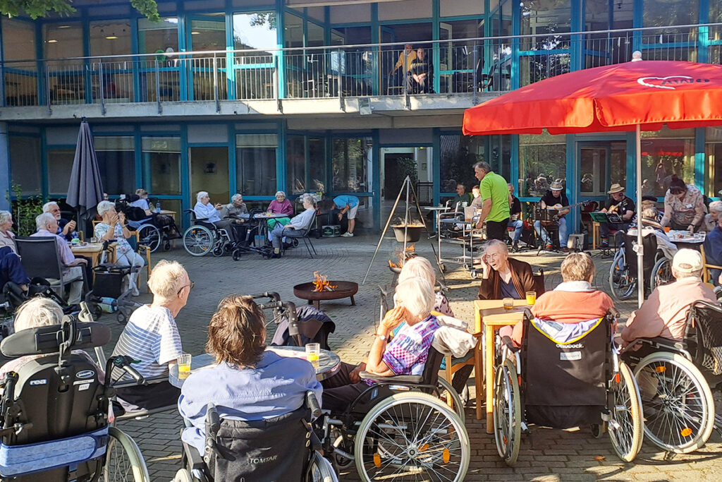 &Auml;ltere Menschen in Rollst&uuml;hlen sitzen im Freien bei einer Veranstaltung vor einem Geb&auml;ude.