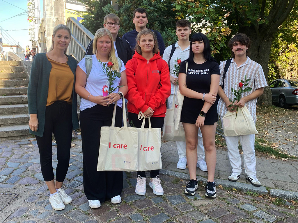 Eine Gruppe von acht Personen steht zusammen auf einem Kopfsteinpflasterweg vor einer Treppe. Einige der Personen halten Taschen mit der Aufschrift 'I care' und Rosen in den Händen.