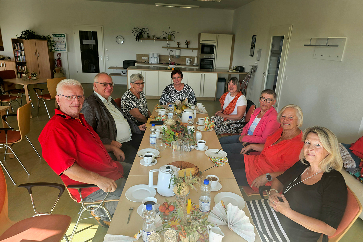 Gruppe von Menschen sitzt um einen gedeckten Tisch in einem Gemeinschaftsraum.