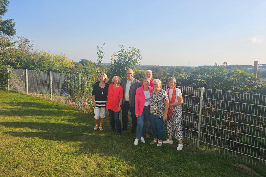 Eine Gruppe von sieben Personen steht auf einer Wiese vor einem Zaun und B&uuml;schen, l&auml;chelnd in die Kamera.
