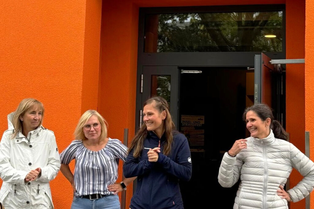 Vier lächelnde Frauen vor einem orangefarbenen Gebäude.