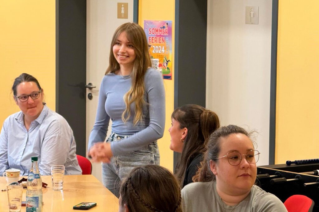 Eine Gruppe von Menschen sitzt an einem Tisch in einem Raum mit gelben W&auml;nden. Eine junge Frau steht und l&auml;chelt, w&auml;hrend andere Personen anwesend sind.