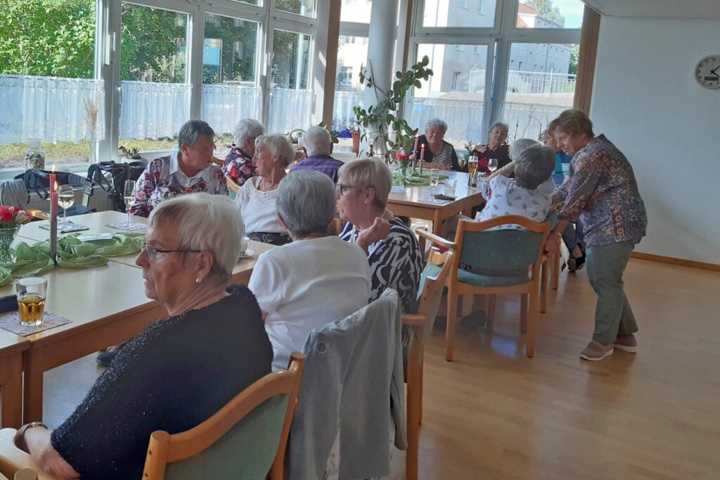 Eine Gruppe &auml;lterer Menschen sitzt an Tischen in einem hellen Raum mit gro&szlig;en Fenstern.