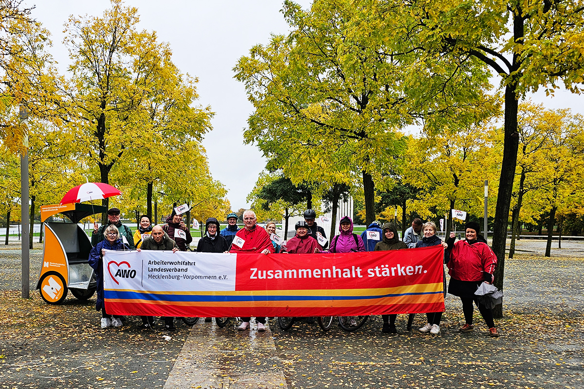 Eine Gruppe von Menschen hält ein Banner mit dem Logo der AWO und der Aufschrift ‚Zusammenhalt stärken‘ in einem herbstlichen Park.