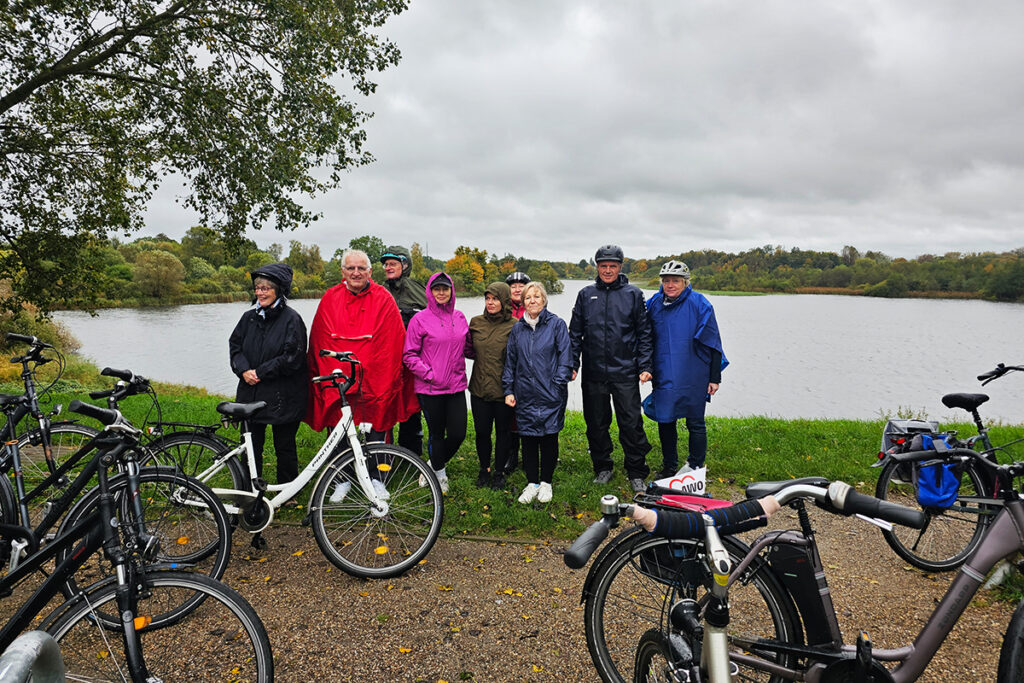 Eine Gruppe von Menschen in Regenm&auml;nteln steht mit Fahrr&auml;dern vor einem See.