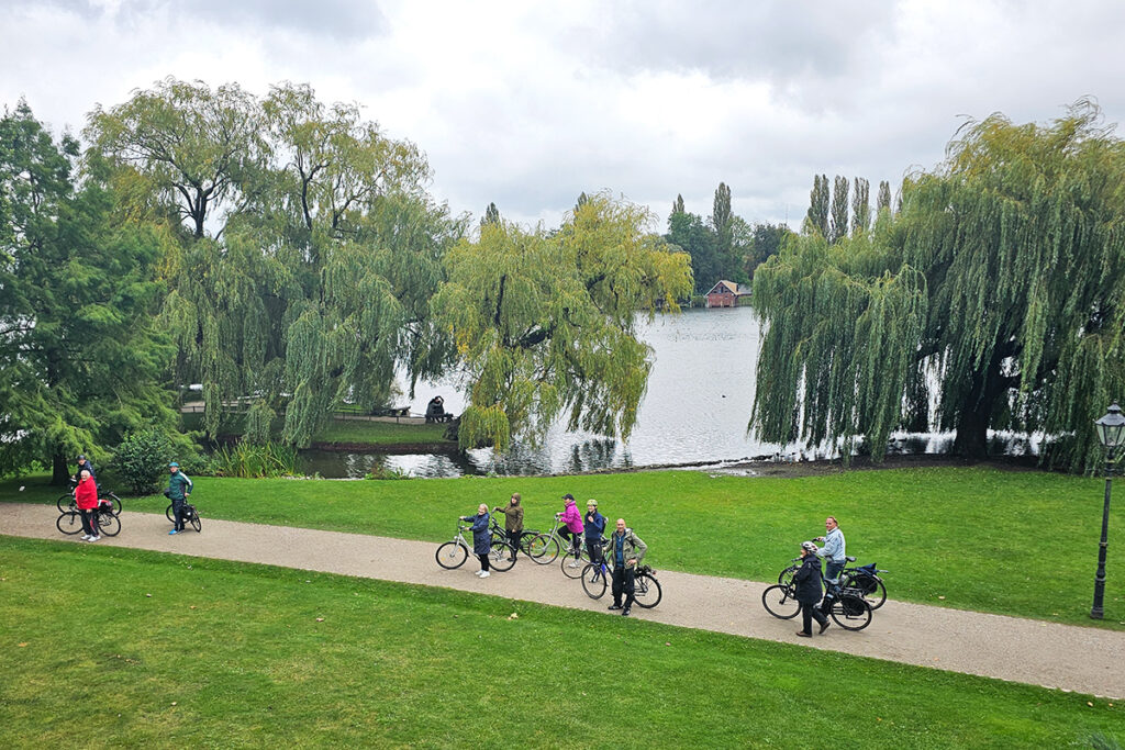 Eine Gruppe von Radfahrern auf einem Parkweg neben einem See mit B&auml;umen.