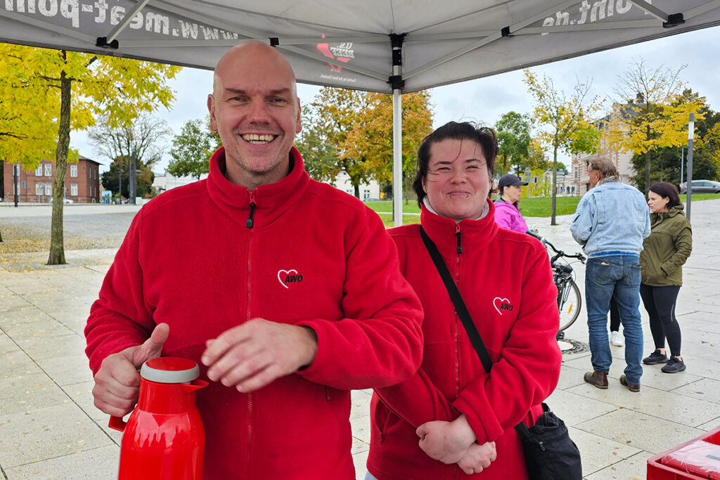 Zwei Personen in roten AWO-Jacken stehen unter einem Pavillon und l&auml;cheln in die Kamera.