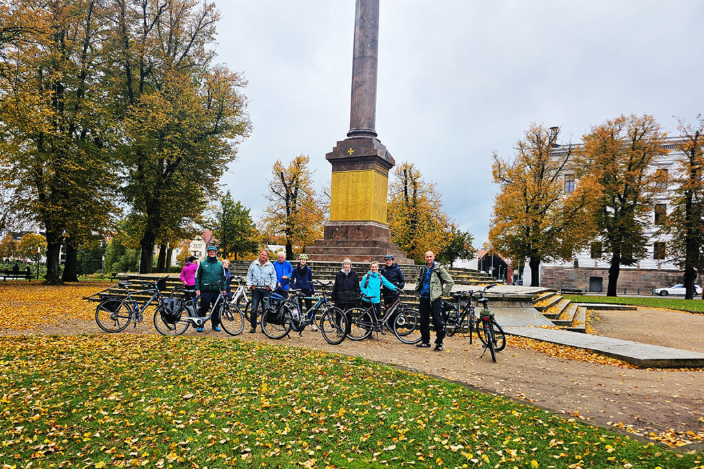Gruppe von Menschen mit Fahrr&auml;dern vor einer hohen S&auml;ule im Herbstpark