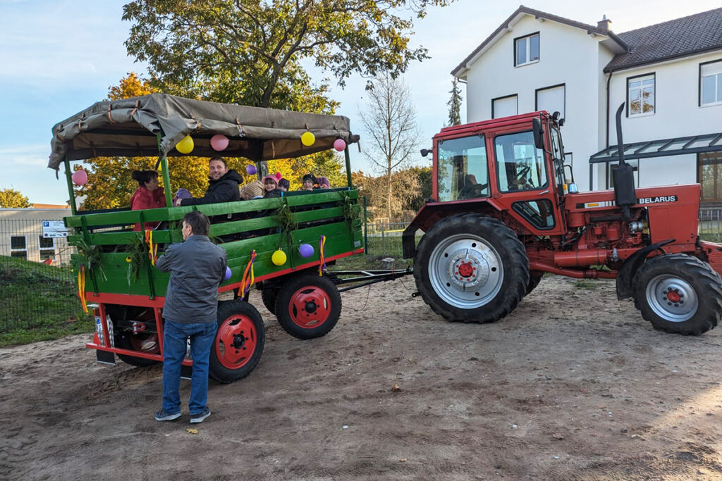 Ein roter Belarus-Traktor zieht einen gr&uuml;n dekorierten Anh&auml;nger mit Menschen darauf.