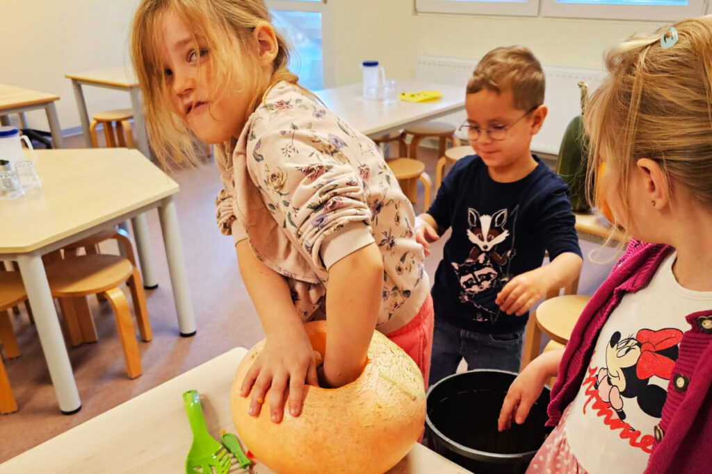 Kinder spielen mit einem K&uuml;rbis in einem Raum mit Tischen und Hockern.