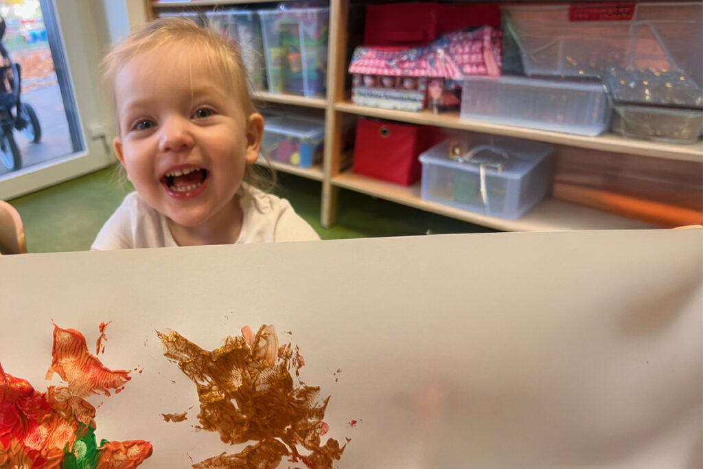 Ein l&auml;chelndes Kleinkind in einer Kita mit einem Gem&auml;lde in der Hand.