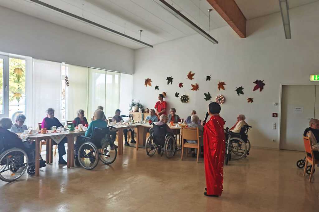 Menschen in einem Gemeinschaftsraum, teilweise mit Rollst&uuml;hlen, an gedeckten Tischen mit herbstlicher Dekoration.