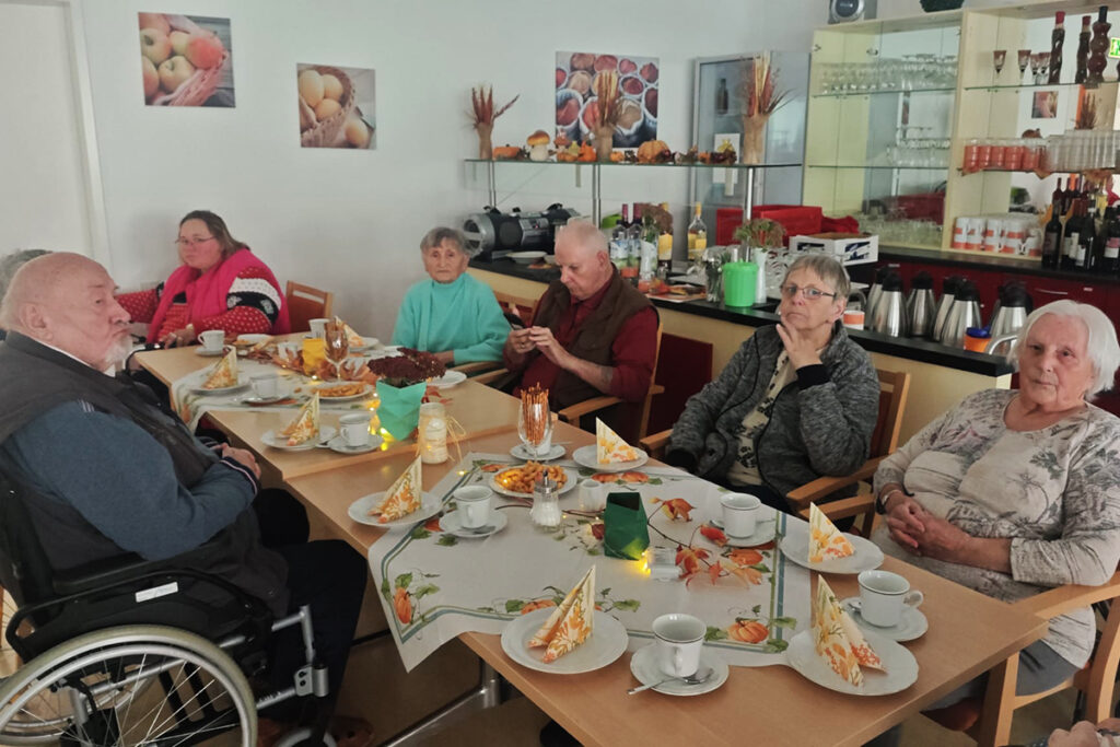 Eine Gruppe von &auml;lteren Menschen sitzt um einen gedeckten Tisch in einem Gemeinschaftsraum.