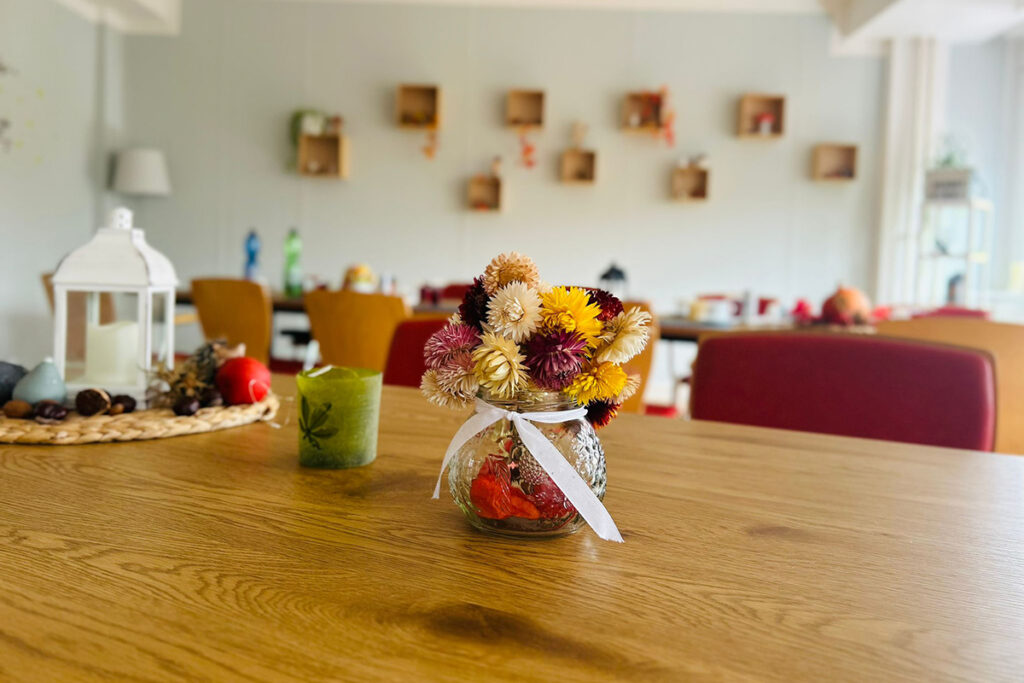 Ein gem&uuml;tlicher Raum mit einem Holztisch, auf dem ein Glasgef&auml;&szlig; mit Trockenblumen steht.