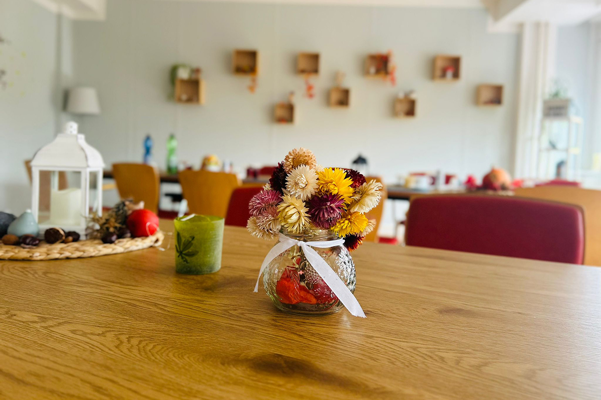 Ein gemütlicher Raum mit einem Holztisch, auf dem ein Glasgefäß mit Trockenblumen steht.