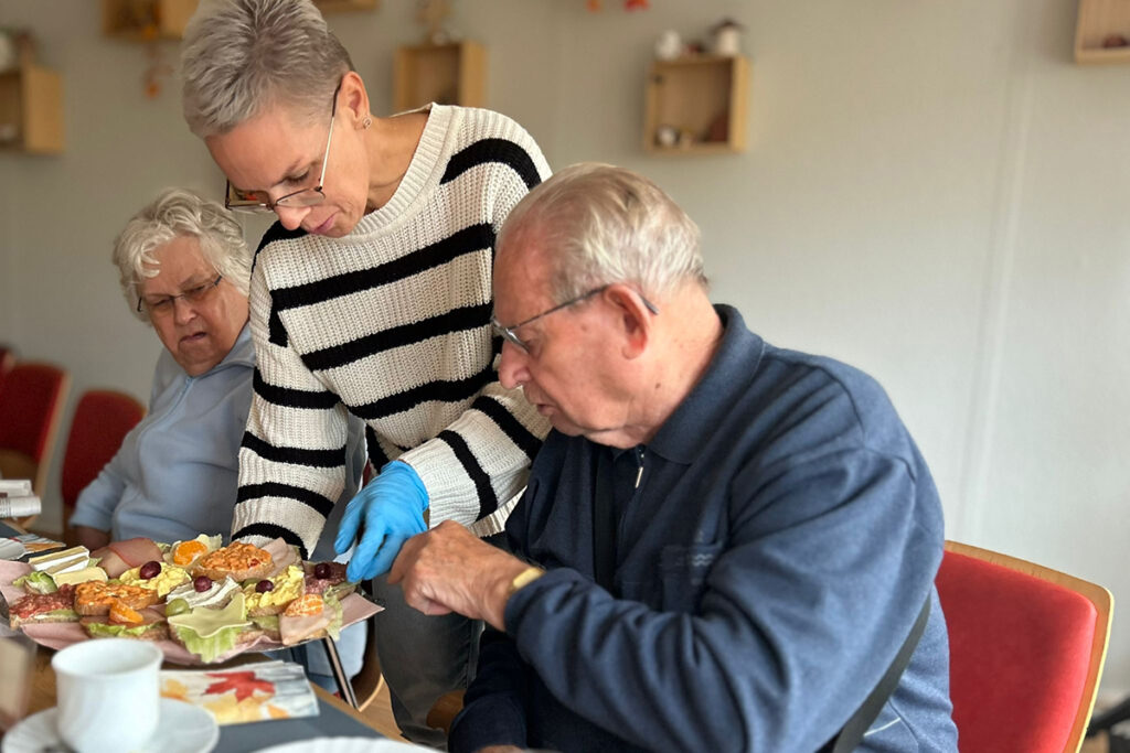 Eine Person in einem gestreiften Pullover hilft einem &auml;lteren Mann am Tisch, w&auml;hrend eine weitere &auml;ltere Frau zuschaut.