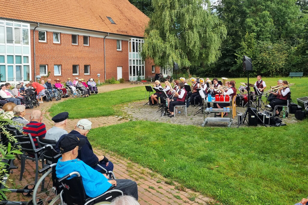 Ein Konzert im Freien f&uuml;r Senioren, aufgef&uuml;hrt von einer Blaskapelle vor einem roten Backsteingeb&auml;ude.