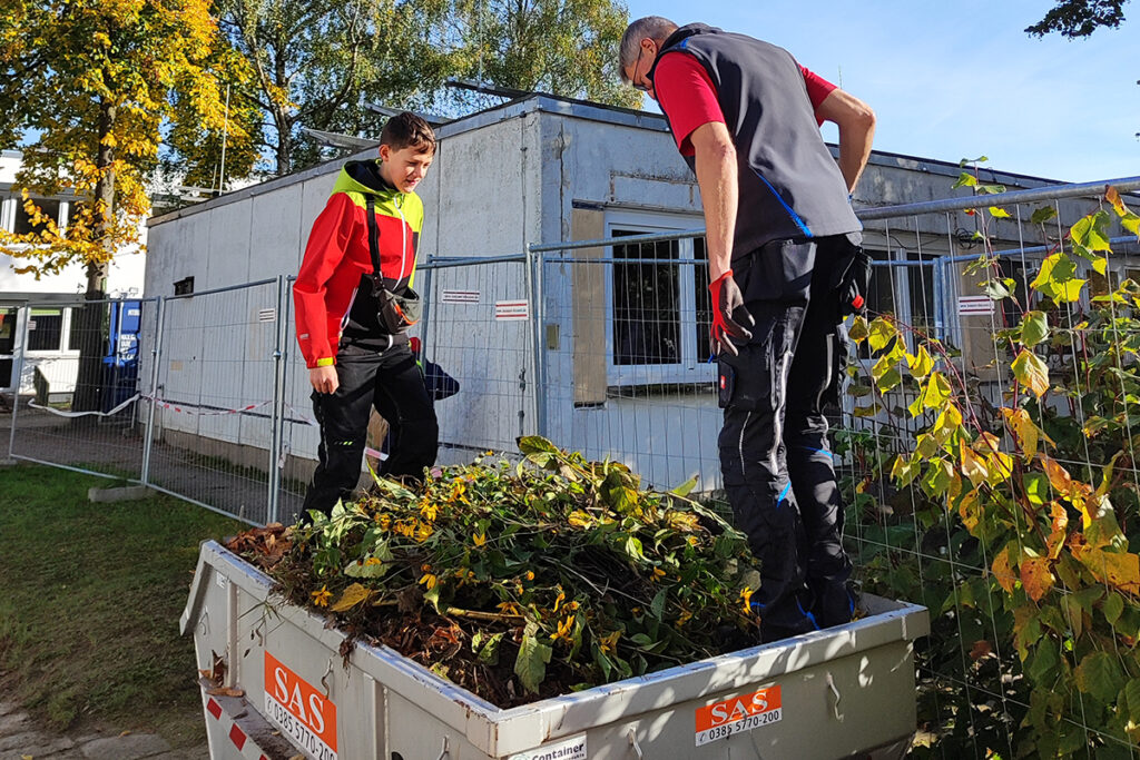Zwei Personen stehen auf einem Anh&auml;nger voller Gartenabf&auml;lle vor einem Geb&auml;ude.