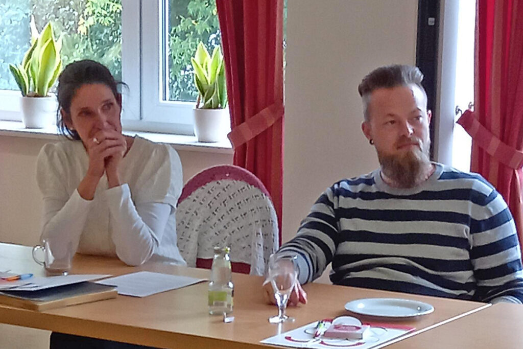 Zwei Personen sitzen an einem Tisch in einem Raum mit roten Vorh&auml;ngen.