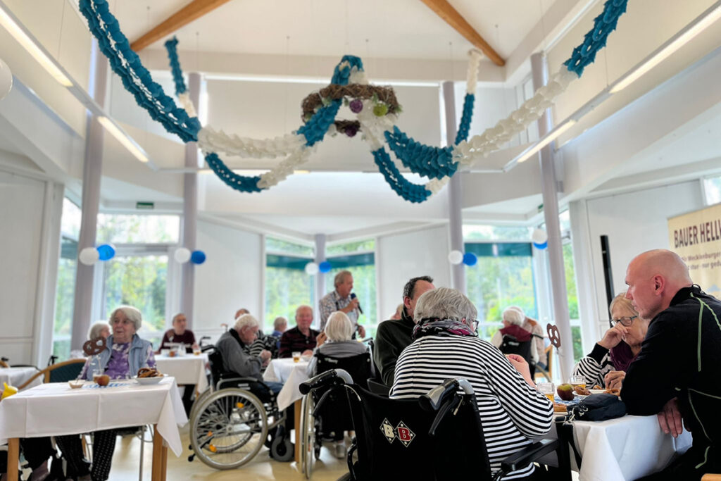 Foto von Menschen in einem Gemeinschaftsraum, einige in Rollst&uuml;hlen, bei einer Veranstaltung mit blau-wei&szlig;er Dekoration.