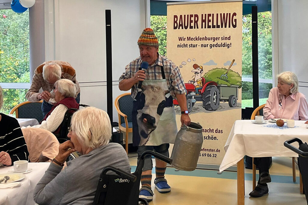 Mann mit Milchkannen-Kost&uuml;m h&auml;lt Mikrofon vor Seniorenpublikum