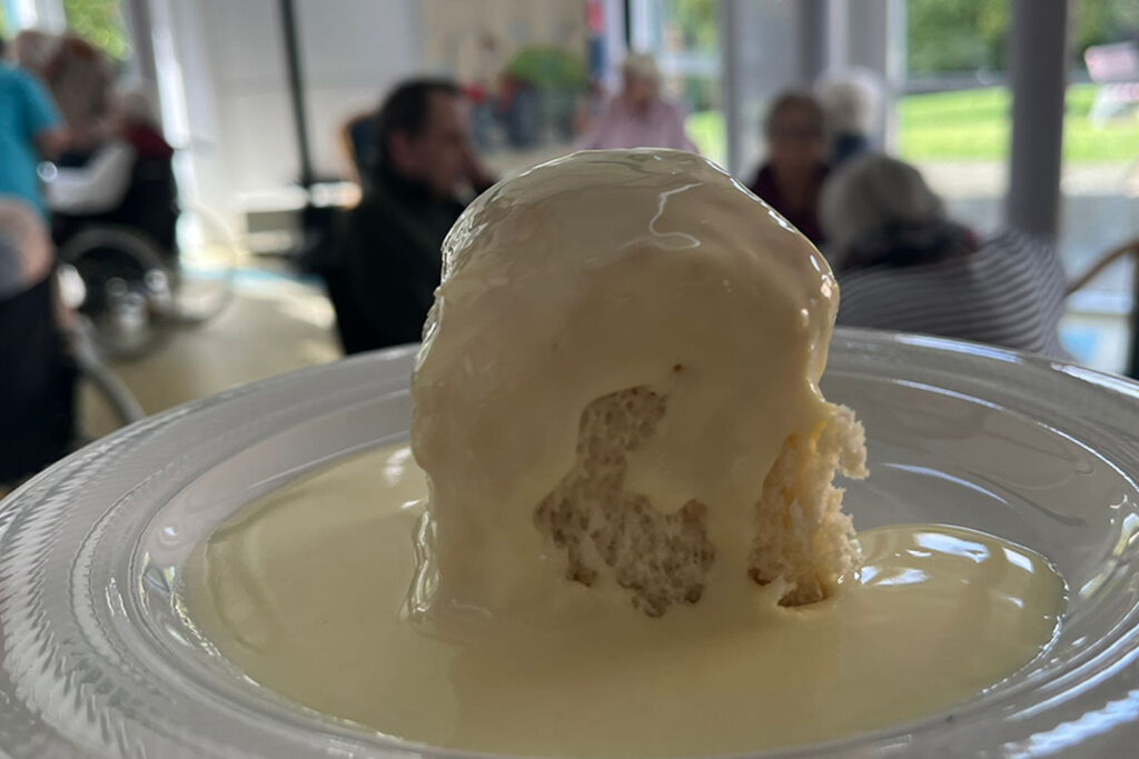 Ein Dessert mit Vanillesauce im Vordergrund, Personen im Hintergrund unscharf dargestellt.