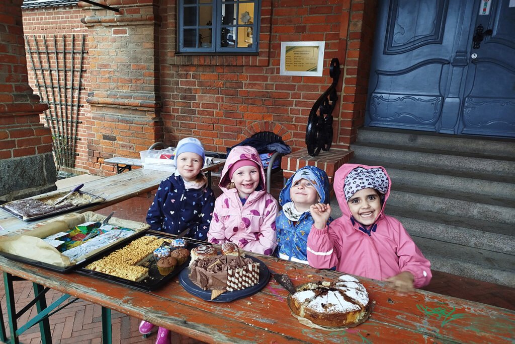 Vier Kinder sitzen an einem Tisch mit Kuchen vor einem Backsteingeb&auml;ude.